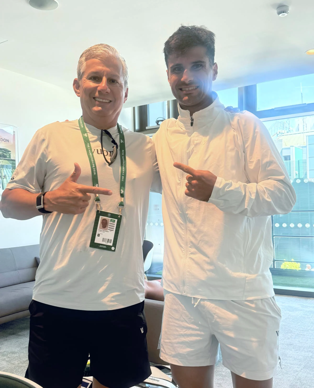 Rob Borrego and Marcos Giron smiling and pointing at each other's Wimbledon credentials in the players' lounge